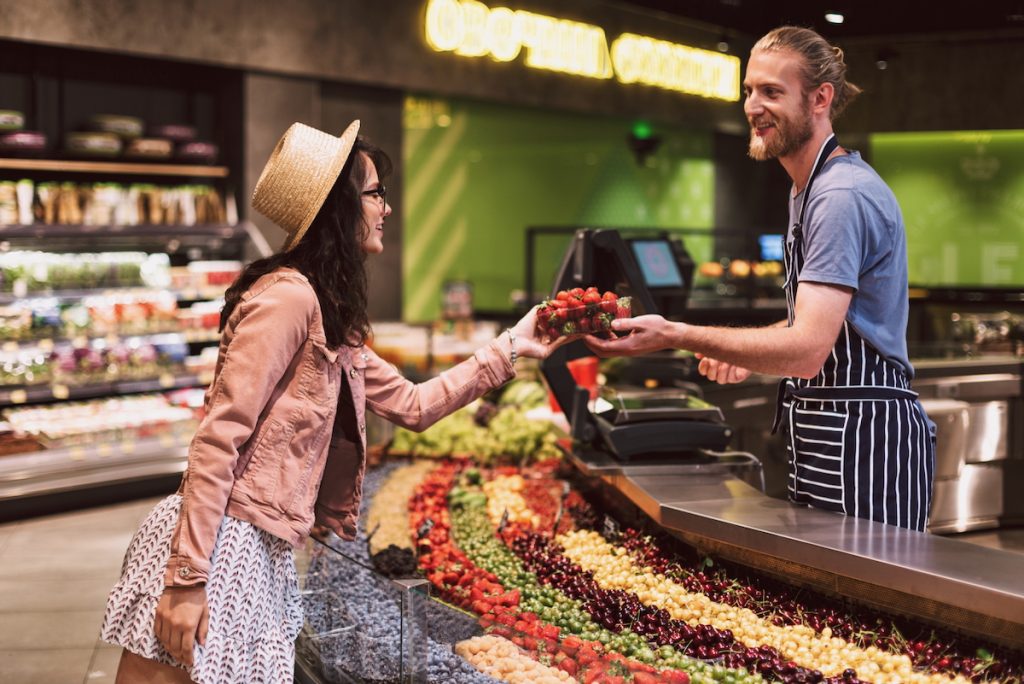 Retail and wholesale - young smiling seller in apron behind counter happily giving strawberries to customer in supermarket
